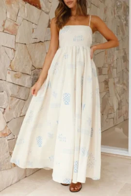 Front view of a woman wearing a cream linen maxi dress with blue Mediterranean graphic prints, sun motifs, tile patterns, and "Ciao Bella" text across the chest.