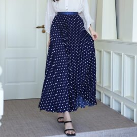 Model wearing a high waisted navy blue pleated midi skirt with large white polka dots paired with a white long-sleeve blouse.